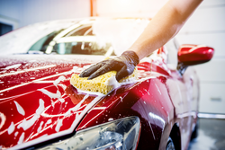 A red car covered in soapy water being washed by a man with a sponge