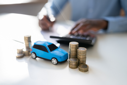 A blurred photo of a man using a calculator and making notes with a pile of coins and a toy car in front of him